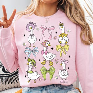 Puede incluir: Sudadera rosa con un estampado gr&aacute;fico de gansos con sombreros y sosteniendo lazos. Los gansos est&aacute;n en varias poses y est&aacute;n rodeados de flores y estrellas.