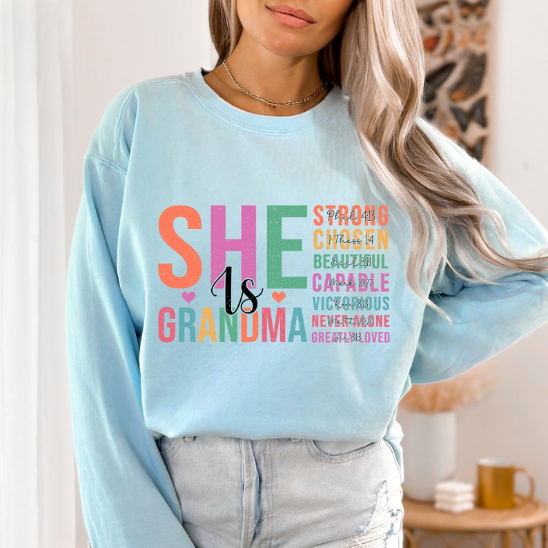 May include: A light blue sweatshirt with the text "She is Grandma" in colorful letters. The text is surrounded by other words in smaller font, including "Strong", "Chosen", "Beautiful", "Capable", "Victorious", "Never Alone", and "Greatly Loved".