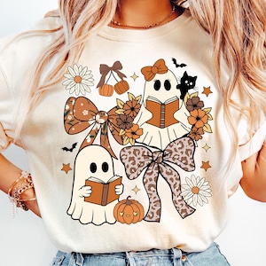 Op de afbeelding: Cr&egrave;mekleurig grafisch T-shirt met grillige Halloween-illustraties. Twee spookfiguren lezen boeken, omringd door strikken, bloemen, pompoenen, sterren en een zwarte kat. Het ontwerp bevat tinten bruin, oranje en cr&egrave;me.