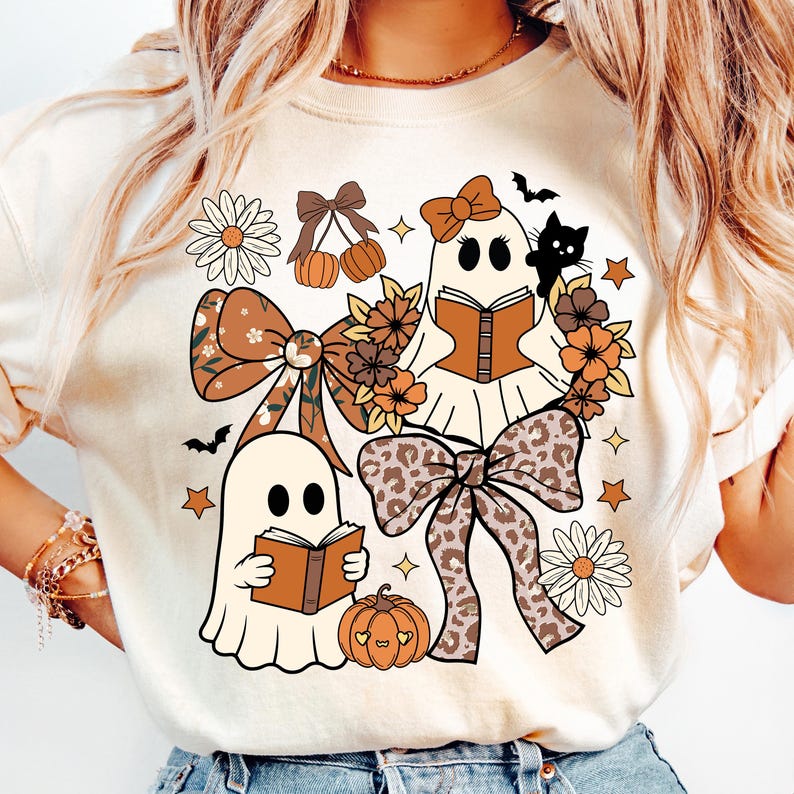 Op de afbeelding: Cr&egrave;mekleurig grafisch T-shirt met grillige Halloween-illustraties. Twee spookfiguren lezen boeken, omringd door strikken, bloemen, pompoenen, sterren en een zwarte kat. Het ontwerp bevat tinten bruin, oranje en cr&egrave;me.