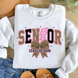 Pu&ograve; includere: Una felpa grigio chiaro con la scritta "SENIOR" in un font leopardato, un fulmine rosa e un fiocco leopardato. Il testo "CLASS OF 2026" &egrave; sotto il fiocco. La felpa &egrave; del marchio Gildan.