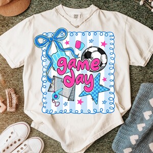 May include: Cream-colored t-shirt featuring a colorful "Game Day" graphic. The design includes a soccer ball, stars, a bow, and other sports-themed elements in blue and pink. The shirt is made of a soft, comfortable material.