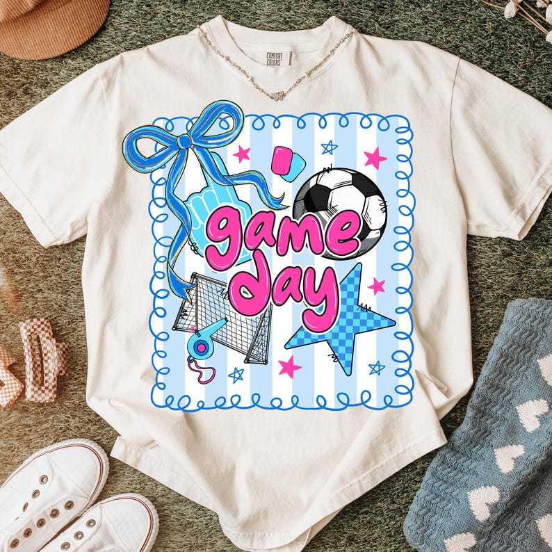 May include: Cream-colored t-shirt featuring a colorful "Game Day" graphic. The design includes a soccer ball, stars, a bow, and other sports-themed elements in blue and pink. The shirt is made of a soft, comfortable material.