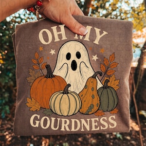 May include: A brown t-shirt featuring a graphic of a white ghost surrounded by orange, yellow, and green pumpkins and gourds, with autumn leaves and stars. The text reads OH MY GOURDNESS in a cream-colored font above and below the illustration.