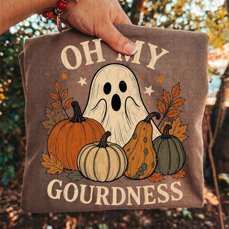 May include: A brown t-shirt featuring a graphic of a white ghost surrounded by orange, yellow, and green pumpkins and gourds, with autumn leaves and stars. The text reads OH MY GOURDNESS in a cream-colored font above and below the illustration.