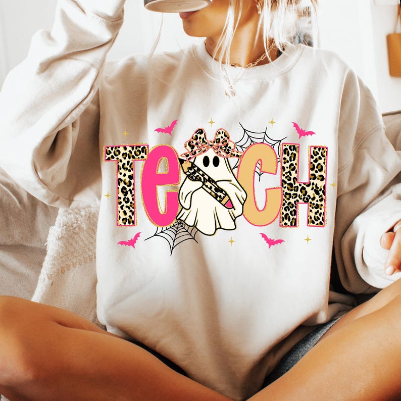 May include: Cream-colored sweatshirt with the word "TEACH" in a Halloween-themed design. The letters "T" and "H" have a leopard print pattern. The "E" is pink, and the "C" is orange. A ghost wearing a bow and holding a pencil is in the center.