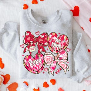 Puede incluir: Una sudadera gris claro con un dise&ntilde;o de San Valent&iacute;n. El dise&ntilde;o incluye lazos rojos, caramelos en forma de coraz&oacute;n con las frases "Be Mine" y "XOXO", y peque&ntilde;os corazones. Los lazos tienen un estampado de lunares y corazones.