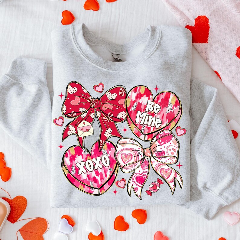 Puede incluir: Una sudadera gris claro con un dise&ntilde;o de San Valent&iacute;n. El dise&ntilde;o incluye lazos rojos, caramelos en forma de coraz&oacute;n con las frases "Be Mine" y "XOXO", y peque&ntilde;os corazones. Los lazos tienen un estampado de lunares y corazones.