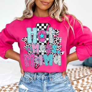 May include: Pink sweatshirt with a graphic print of a lightning bolt with a checkered pattern and the text "Hot Mess Mama" in pink, blue, and black with animal print accents.