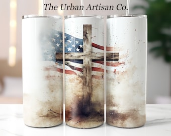 May include: A white tumbler with a watercolor design featuring the American flag and a wooden cross. The design has a distressed, vintage look with brown and red accents. The text "The Urban Artisan Co." is at the top.