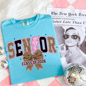 Pu&ograve; includere: Una maglietta azzurra con la scritta "SENIOR" in un carattere leopardato con contorni neri. Sono presenti anche un fulmine rosa e un fiocco leopardato. Il testo "CLASS OF 2026" &egrave; sotto il fiocco. Un ritaglio di giornale &egrave; sullo sfondo.