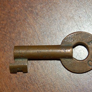 May include: A vintage brass key with a circular head and a rectangular bit. The head of the key has the letters "PTCO" stamped on it. The key is a warm, antique brass color.