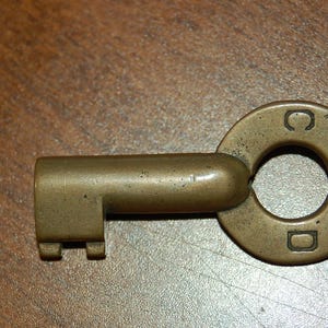 May include: A vintage brass key with a circular head and a rectangular key bit. The head is stamped with the letters "C" and "D" and the number "2744". The key is lying on a brown wooden surface.