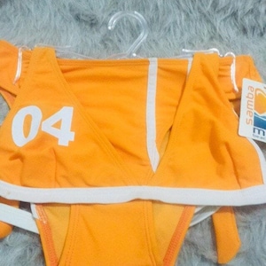 Può includere: Un bikini arancione brillante con bordi bianchi. Il top presenta il numero 04 in un grande carattere bianco sul lato destro. Un'etichetta bianca con la scritta samba è attaccata al lato dello slip del bikini.