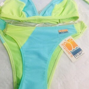 Può includere: Un bikini a due pezzi con blocchi di colore verde lime brillante e azzurro cielo. Il top presenta coppe triangolari e lo slip è a taglio alto. Un'etichetta rettangolare in metallo con logo è fissata sul fianco, insieme a un cartellino bianco con la scritta Samba mar beachwear.
