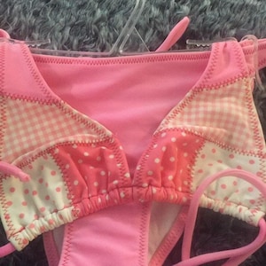 Może przedstawiać: Różowy dół od bikini z patchworkowym wzorem, składający się z naprzemiennych paneli z gładkiej różowej tkaniny, biało-różowej kratki gingham oraz białych kropek na różowym tle. Strój posiada wiązania po bokach i widoczne szwy zygzakowate.