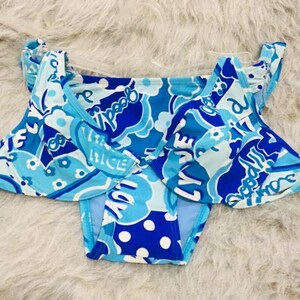 Può includere: Un bikini a due pezzi con un vivace motivo blu e azzurro, pois bianchi, disegni floreali e la scritta "lovely" e "goodie" ripetuta sul tessuto. Il top presenta coppe con ferretto e spalline sottili.