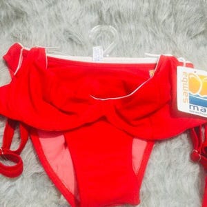 May include: A bright red bikini bottom featuring a high-waisted design with a ruffled overlay across the front. The garment includes a white trim along the top edge and side ties with metal hardware. A white tag labeled samba beachwear is attached.