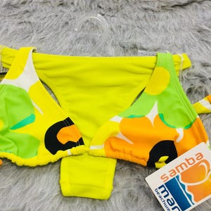 Puede incluir: Un conjunto de bikini amarillo brillante con un llamativo estampado floral en tonos verde lima, naranja y negro. La parte superior tiene copas triangulares y la parte inferior es de corte clásico. Incluye una etiqueta blanca con el texto samba mar beachwear.
