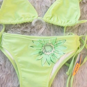 Può includere: Un bikini a due pezzi color verde lime composto da un top a triangolo e uno slip abbinato con laccetti laterali. Lo slip del bikini è decorato con una grande grafica a forma di girasole dipinta a mano nei toni del verde chiaro e bianco.