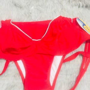 Puede incluir: Un top de bikini rojo brillante con un volante superpuesto y un fino borde blanco a lo largo del escote. La prenda de baño incluye lazos laterales y un forro interior blanco, presentada en una percha de plástico transparente con etiqueta.
