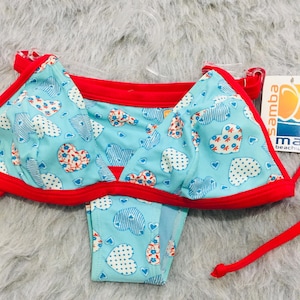 Può includere: Un set bikini azzurro con un motivo ripetuto di cuori bianchi e rossi con dettagli floreali e a righe. Il top e lo slip del bikini sono rifiniti con bordi in tessuto rosso brillante e includono sottili laccetti rossi sui lati.