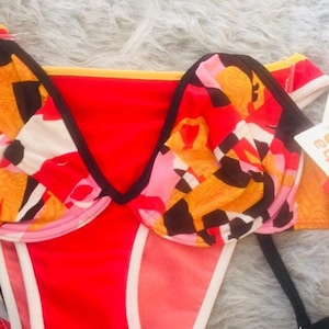 Può includere: Un top bikini con un vivace motivo astratto nei toni del rosso, arancione, rosa, nero e bianco. Il capo presenta un design strutturato con ferretto, bordi neri lungo le coppe e un dettaglio a fascia gialla sul bordo superiore.