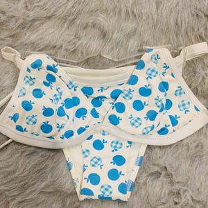 Può includere: Un set bikini a due pezzi con base bianca e un motivo ripetuto di mele blu a tinta unita e mele a quadretti vichy blu. Il top è dotato di ferretto e spalline sottili, mentre lo slip è un classico modello da bikini.