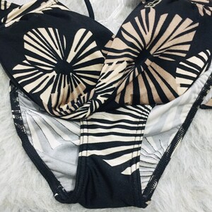 May include: A two-piece bikini set featuring a black and beige floral pattern. The top has a twisted front detail with thin straps, and the bottom features a matching black and beige striped design. A small white product tag is attached to the top.