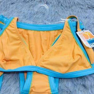 Puede incluir: Un conjunto de bikini de dos piezas en tela naranja brillante con ribetes azul claro vibrantes y detalles en verde lima. La parte superior tiene una silueta triangular y la inferior un corte clásico. Incluye una etiqueta blanca con el texto samba.