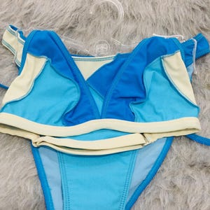 Könnte beinhalten: Ein zweiteiliges Bikini-Set mit einem leuchtend blauen und hellblauen Color-Block-Design und cremefarbenen Abschlüssen. Das Oberteil hat Rüschendetails am Ausschnitt, während das Unterteil seitliche Bänder und einen klassischen Schnitt aufweist.