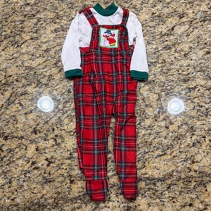 May include: A long-sleeved white shirt with a dark green mock neck collar and cuffs, layered under red, green, and white plaid overalls. The overalls feature a square bib with a central patch depicting a snowman wearing a blue hat and scarf.