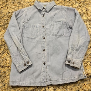 May include: A light blue long-sleeve button-down shirt made of corduroy fabric. The shirt features a standard collar, two chest pockets with flaps, and dark brown buttons down the front placket and on the cuffs of the long sleeves.