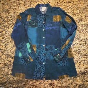May include: A long-sleeved button-down shirt featuring a patchwork design in shades of blue, teal, and gold. The pattern includes abstract shapes, dark blue polka dots, and fine line drawings of foliage. It has a standard collar and buttons.