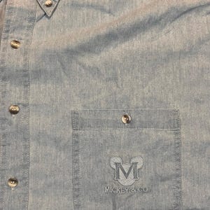 May include: Light blue button-down shirt with a chest pocket. The pocket features an embroidered "M" logo with "MICKEY & CO" text. The shirt has several buttons and a classic collar.