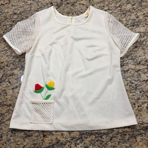 Op de afbeelding: Een crèmekleurige top met korte mouwen, witte mesh mouwen en een bijpassende mesh zak aan de linkeronderkant. De zak bevat twee stoffen bloemen, één rood en één geel, met groene stelen en bladeren die aan het kledingstuk zijn bevestigd.