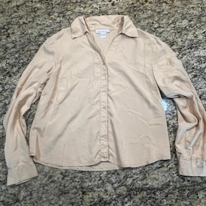May include: A light beige button-down shirt with long sleeves. The shirt has a collared neckline and a series of small buttons down the front. The fabric appears to be a lightweight material.