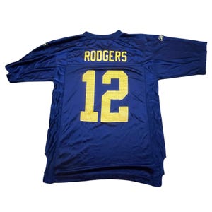 Puede incluir: Una camiseta de fútbol azul marino con el nombre "RODGERS" y el número "12" en amarillo. La camiseta está hecha de un material brillante y atlético y tiene mangas cortas.