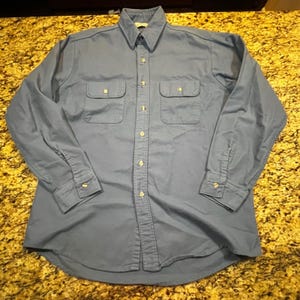 Puede incluir: Una camisa azul de manga larga con botones y dos bolsillos en el pecho. La camisa tiene cuello y puños abotonados. La tela parece ser de estilo trabajo duradero. La camisa está tendida sobre una superficie texturizada.