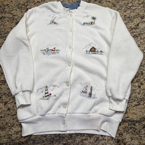 May include: A white long-sleeve button-down cardigan featuring a textured fabric and embroidered nautical scenes. The embroidery includes sailboats, a lighthouse, a beach house, and palm trees arranged in a grid pattern across the front.