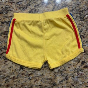 May include: Bright yellow shorts featuring a solid yellow main body with a thick red vertical stripe flanked by a thin blue stripe running down each side. The shorts have a simple elasticized waistband and a hemmed leg opening design.