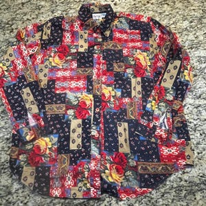 May include: A long-sleeved button-down shirt with a patchwork design. The shirt features floral and geometric patterns in red, yellow, navy, and tan. It has a collared neckline and the label reads "Jacqueline de Yahia".