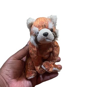 May include: A plush red panda toy with orange and white fur, black eyes, and white whiskers. The stuffed animal is sitting upright, with its paws in front. The toy is designed to resemble a red panda.
