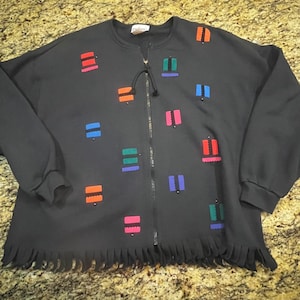 May include: Black zip-up sweatshirt with colourful geometric accents. The sweatshirt features a black zipper, a drawstring, and a fringe detail at the bottom. The accents are in shades of red, orange, blue, green, and pink.