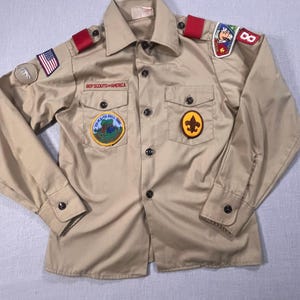 May include: A tan Boy Scouts of America shirt with various patches, including a US flag and a fleur-de-lis. The shirt features long sleeves, two chest pockets, and red shoulder epaulettes.
