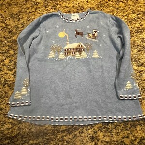 Könnte beinhalten: Ein hellblauer Langarmpullover mit einer gestickten Winterszene, die ein Haus, einen gelben Mond und einen von einem Rentier gezogenen Schlitten zeigt. Der Halsausschnitt, die Ärmelbündchen und der Saum sind mit einem braun-weißen Karomuster versehen.