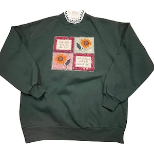 Puede incluir: Sudadera verde oscuro con cuello a cuadros blancos y negros. La parte delantera presenta cuatro cuadrados bordados con girasoles y las frases "Keep your face to the sun" y "And shadows will fall behind you."