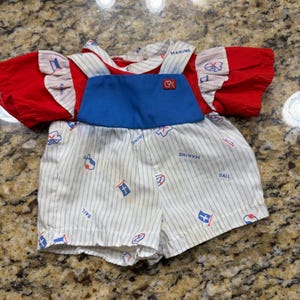 May include: A doll outfit consisting of a red short-sleeved shirt with ruffled shoulders and white pinstriped fabric, paired with white pinstriped shorts featuring a blue bib front, nautical icons, and the text MARINE and SAIL with a CPK logo patch.