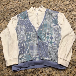May include: A long-sleeve pullover shirt featuring a mock neck with a three-button placket. The front panel displays a blue and white patchwork pattern with various geometric and floral designs, while the sleeves and collar are solid cream.