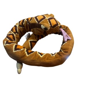 May include: A plush snake toy in shades of brown, orange, and tan, with a diamond pattern. The snake has a small head with a pink tongue sticking out. The toy is curled into a loop.